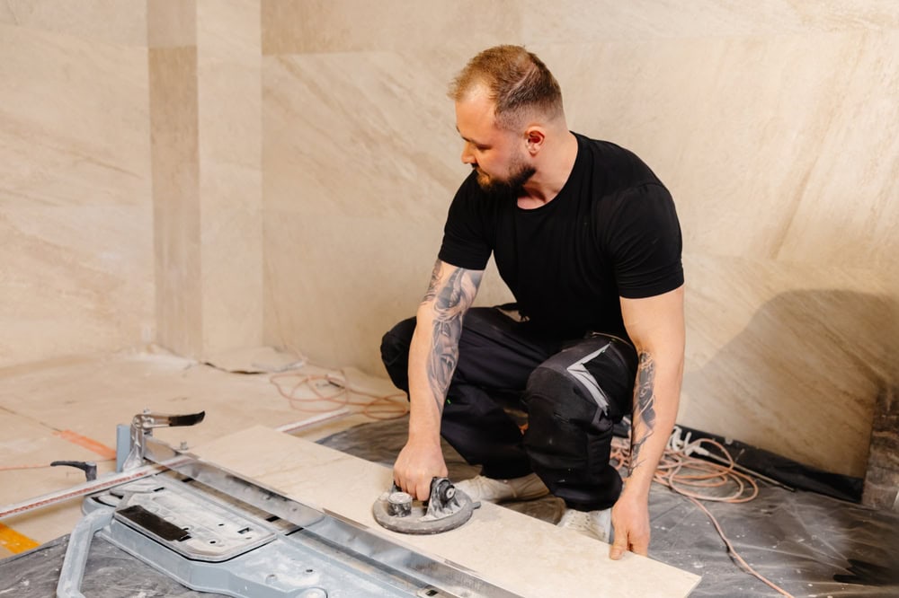 A man with tattoos and a beard wearing a black shirt is kneeling on the floor, using a tool to lift a large tile while working on Flooring Installation in Wake County, NC and Horry County.