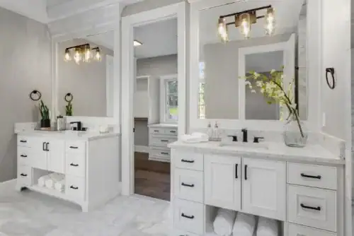 Modern bathroom with two white vanities, large mirrors, black fixtures, and marble floor from Flooring Installation Wake County, NC. Decorative lights, potted plants, and rolled towels on shelves add style. A doorway shows another room with similar decor.