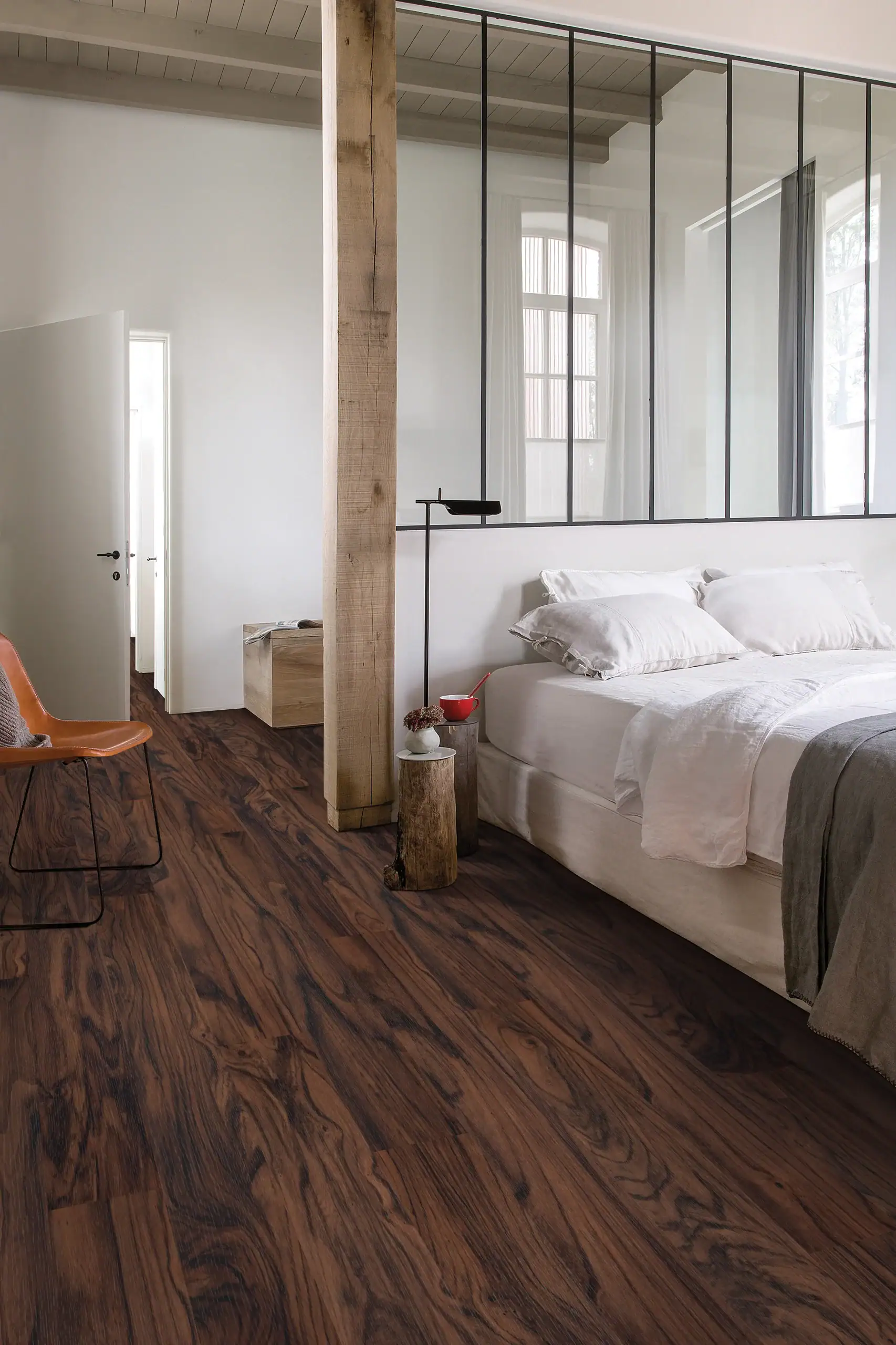 A modern bedroom with white walls, large windows, and a glass partition showcases elegant dark wood patterns—perfect inspiration for Flooring Installation Wake County, NC and Horry County, SC. An orange chair and side tables complete the look.