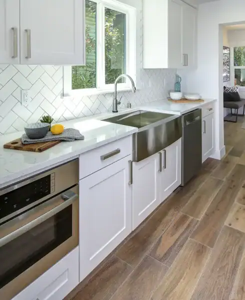 A modern kitchen with white cabinets, stainless steel appliances, and wood-look flooring—perfect for those seeking quality Flooring Installation in Wake County, NC and Horry County. Natural light pours in through a large window above the countertop.