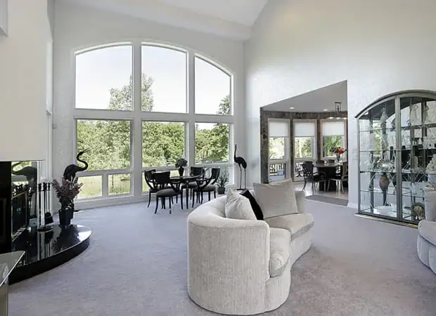 'n Helder sitkamer met groot geboë vensters, moderne roomkleurige sofas en ligte matte van die boonste vloerinstallasie Wake County, SC, NC en Horry County; met 'n swart kaggel, glasuitstalkas en 'n eetarea met uitsig oor groen buitelug.