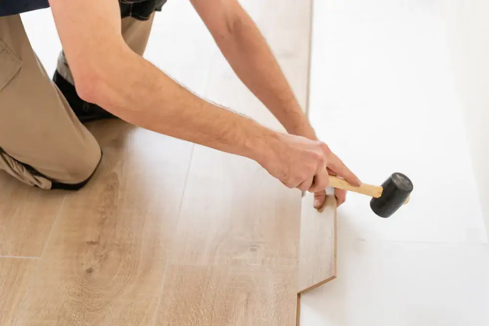 'n Persoon kniel op die vloer met 'n rubberhamer om ligte laminaatvloere met 'n houtagtige voorkoms te installeer, en pas 'n plank met 'n sigsagrand langs 'n wit muur &ndash; perfek vir vloerinstallasie in Wake County, NC en Horry County.