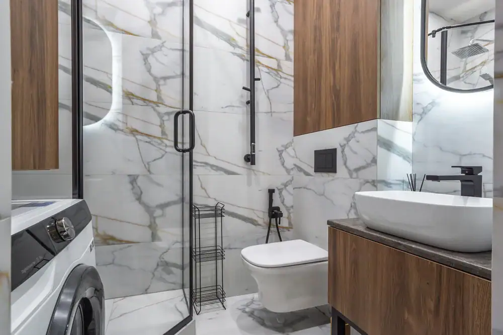 Moderne badkamer met wit marmermure, houtkaste en 'n glasstort met 'n swart raam. Met kundige vloerinstallasie in Wake County-styl, is die ruimte helder, netjies georganiseerd en sluit 'n muurhangende toilet, wasbak en wasmasjien in.