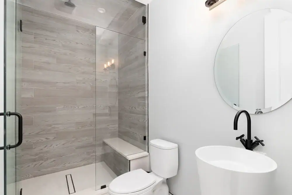 Moderne badkamer met 'n glas-ingehulde stort met ligte houtpatroonte&euml;ls en stylvolle vloerinstallasie&mdash;bedien Wake County, SC, NC en Horry County&mdash;met 'n ingeboude bankie, wit toilet, voetstukwasbak met swart kraan en ronde spie&euml;l teen 'n wit muur.