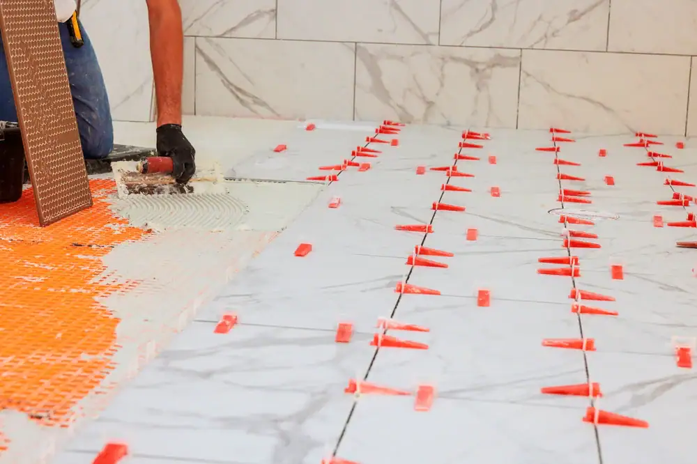 'n Werker voltooi vloerinstallasie in Wake County, NC en Horry County, waar hy kleefmiddel vir groot marmerteëls versprei. Oranje spasieerders en gelykmaakklemme verseker egalige plasing, met marmermuurteëls sigbaar in die agtergrond.